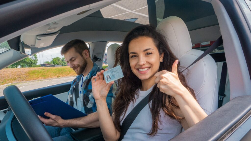 Lächelnde junge Frau mit Führerschein, die Daumen nach oben zeigt, und Fahrschullehrer mit Klemmbrett im Auto. Fahrprüfung, Fahrkurse, Prüfungskonzept