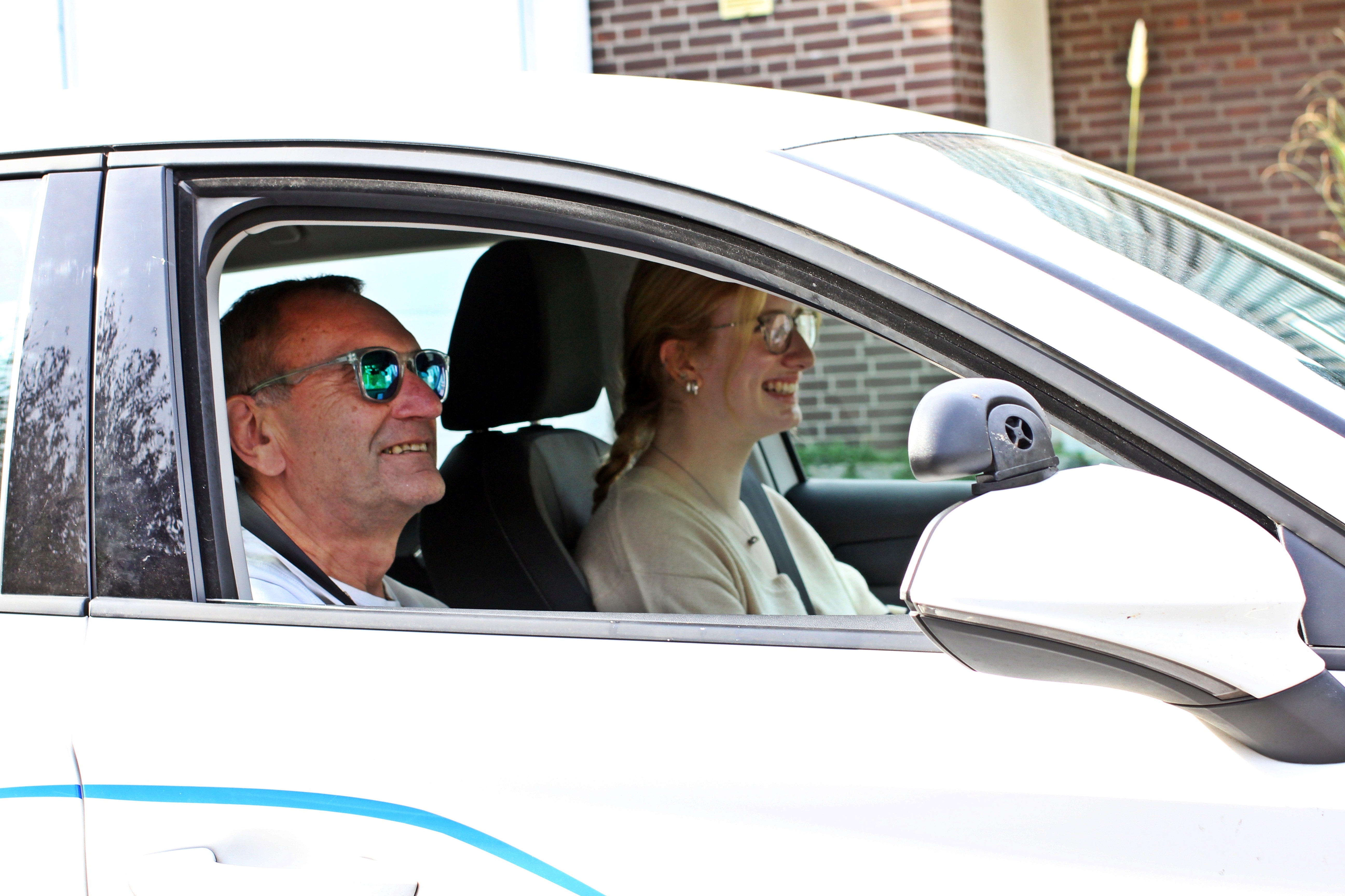 Ein Mann und eine Frau lachen in einem weißen Auto. Mann trägt Sonnenbrille.